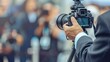 © Jiraphat - A photographer in a suit holds a camera on a tripod, focused on capturing an engaging event as excited attendees surround the area