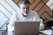 © opolja - Smiling mature business man executive wearing shirt sitting at desk using laptop. Happy busy professional middle aged businessman investor working on computer looking away in office