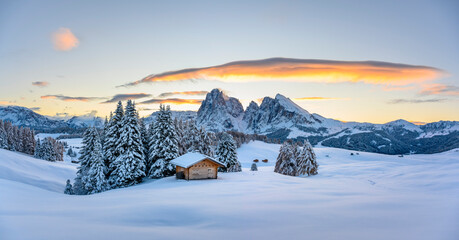 Panoramic view on Alpe di Siusi meadow with small wooden log cabin and first snow. Snowy hills with orange larch and Sassolungo and Langkofel mountains group. Seiser Alm, Dolomites, Italy.