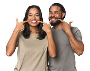 Wall Mural - Young Latino couple in studio smiles, pointing fingers at mouth.