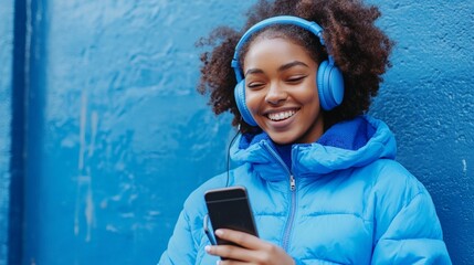 Wall Mural - Joyful young woman with curly hair enjoying music on blue headphones while using smartphone, dressed in vibrant blue jacket against matching wall.