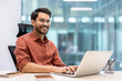 © Liubomir - Smiling man wearing glasses and an orange shirt working on laptop in modern office. Phone and notebook on desk. Bright and professional workspace with glass walls.