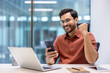 © Liubomir - Excited businessman celebrating success at work using smartphone. Young professional sitting at office desk with laptop, phone, and notebook, showing joyful expression and happiness.