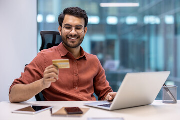 Wall Mural - Smiling man using laptop for online shopping holding credit card. Modern office setting with phone and tablet on desk. Concept of e-commerce, technology, and finance.