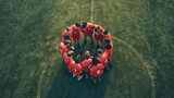 Aerial view of a soccer team huddled in a circle on a green field, wearing red uniforms and discussing strategy before the match.