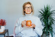 © okrasiuk - Middle aged woman relaxing with pumpkin shaped cup of hot drink in scandy style hygge interior home with fall mood decor. Lady dreaming, enjoy calm mood without stress, well being alone. Cozy autumn