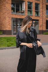 Wall Mural - Elegant young woman looking in her black bag her phone or purse. Traveler style woman wear black leather shirt and bag on the street. Street style, fashion outfit, woman walk on the street