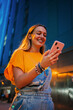 © Jose Calsina - Vertical. one happy young girl using her smartphone to browse the internet while walking on the street at night. Female watching the social media with her cellphone. Girl typing on a mobile phone