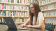 © Pavel - Cute Caucasian brunette with glasses uses laptop to search for student jobs in library, scrolls through newsfeed for research project, exam preparation, higher education online, teachers e-learning