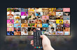 © Prostock-studio - A persons hand holds a remote control and points it towards a large flat screen television. The TV screen displays a variety of colorful, diverse content in a grid format