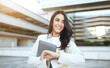 © Prostock-studio - A woman in a white blazer stands in front of a modern building, holding a laptop computer in her left arm. She smiles as she checks her wristwatch with her right hand.