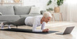 © Prostock-studio - Training At Home. Sporty senior woman doing yoga plank while watching online tutorial on laptop, exercising in living room, free space, panorama