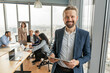 © Prostock-studio - A businessman in a blue suit stands in a modern office, holding a tablet and smiling confidently. He looks directly at the camera. Several other people in business attire are sitting at a table