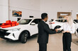 © dikushin - Successful blonde female buyer getting car key and congratulation from black auto dealer male while buying new auto in dealership. Happy young woman car owner during handing over keys at showroom.
