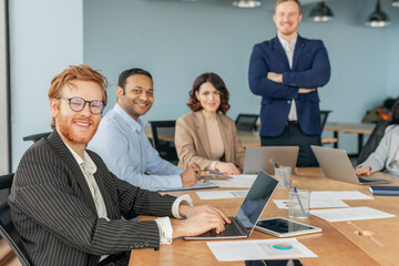 Poster - A group of four business professionals are working together in a modern office setting. One man is seated at a desk and working on a laptop, while another man and a woman are sitting beside him
