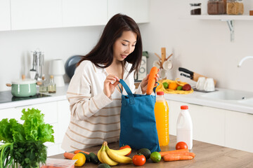 Poster - Young Asian woman unpacking groceries from blue shopper bag onto counter in kitchen