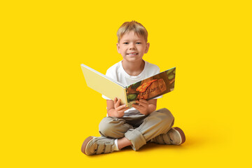 Wall Mural - Cute little boy sitting on floor and reading book on yellow background