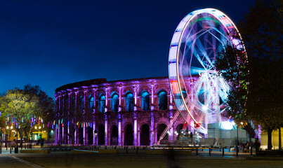 Wall Mural - Spinning ferris wheel with motion blur near illuminated Arena of Nimes, France