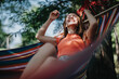 © qunica.com - A joyful woman in a red top relaxes in a colorful hammock under a tree, enjoying a sunny summer day.
