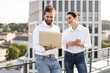 © sofiko14 - Two coworkers smiling while discussing project on rooftop using laptop and phone, wearing casual white shirts and jeans, urban background.