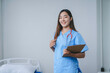 © amnaj - Young female doctor is smiling while holding a clipboard in a hospital room, ready to provide care