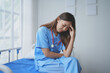 © amnaj - Asian female doctor wearing blue scrubs feeling tired and having a headache while sitting on a hospital bed