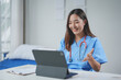 © amnaj - Smiling asian doctor on a video call, embodying modern healthcare. Bright, clean office with stethoscope and paperwork. Joyful, committed to remote patient care