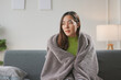 © amnaj - Young woman sitting on a sofa at home, feeling cold and covering herself with a blanket, looking away from the camera