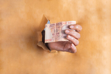 Wall Mural - Woman hand holding Indian 10 Rupee banknote through a hole in brown paper.