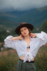 Wall Mural - Fashionable confident woman wearing trendy brown hat, summer white knotted linen shirt, green maxi skirt, posing  among mountains. Outdoor fashion portrait. Copy, empty space for text