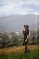 Wall Mural - Fashionable confident woman wearing trendy hat, summer linen shirt, green maxi skirt, carrying black leather bag with fringe, walking among mountains. Outdoor full-length fashion portrait