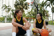 © DragonImages - Two young female basketball players sitting on outdoor court during break, talking and holding basketball, with palm trees and buildings in background