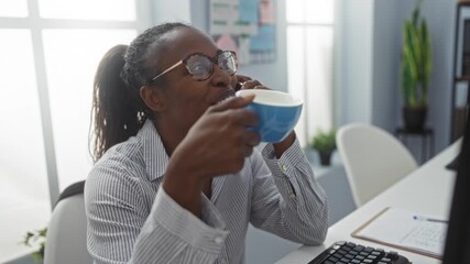 Canvas Print - Young, beautiful, african american woman in an office smiling while drinking from a cup and talking on the phone in a bright, modern workplace setting