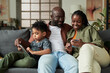 © pressmaster - Young African American couple looking at screen of smartphone while sitting next to their son with touchpad watching online video