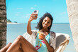 © David Prado/Stocksy - Happy woman takes a selfie at tropical beach with cocktail