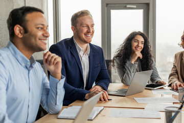 Aufkleber - Four smiling colleagues are sitting around a table, engaged in a conversation during a meeting. The man on the left is listening intently, while the man in the middle is speaking