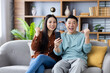 © Liubomir - Cheerful asian couple holding phone celebrating success sitting on couch at home. Couple smiling, displaying excitement and joy for achievement. Shared celebration of happiness and victory