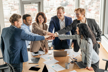 Poster - A diverse group of business professionals stand around a conference table, their hands stacked together in a gesture of teamwork and unity. The office is modern and bright