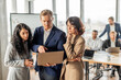 © Prostock-studio - A group of business professionals are gathered around a table in a modern office setting. One man holds a laptop open, pointing at the screen while talking to women standing beside him.