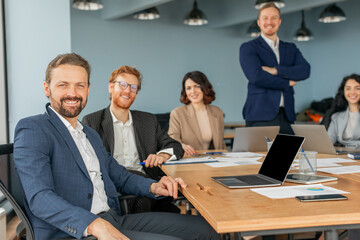 Aufkleber - A group of four business professionals are gathered around a wooden conference table in a modern office. One man sits in the foreground, smiling and looking directly at the camera