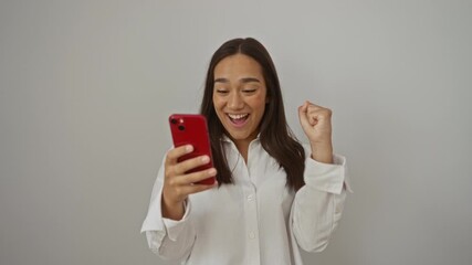 Wall Mural - Woman smiling at phone in bright room celebrating good news with raised hand wearing white blouse using red smartphone.