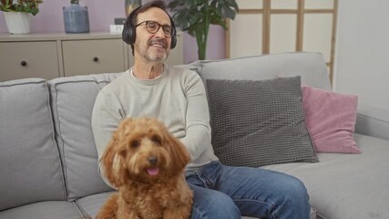 Wall Mural - A smiling middle-aged man with headphones enjoying music on a sofa next to his poodle in a cozy living room.