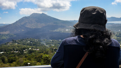 Back view of a female tourist standing on a viewpoint looking at Mount Batur. Volcanic landscape and surrounding greenery are visible under a clear sky, ideal for travel and adventure related content