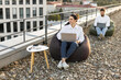 © sofiko14 - Business professionals working on laptops while seated on bean bags on office rooftop. Relaxed work environment embracing outdoor, flexible workspace concept.