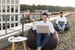 © sofiko14 - Two young professionals working on laptops while sitting on bean bags on rooftop terrace. Surrounded by urban cityscape, enjoying remote working outdoors. Relaxed and productive atmosphere.