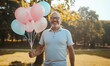 © Nadin Faust - smiling middle-aged man holding pastel blue and pink birthday balloons in a park, celebrating a joyful occasion outdoors
