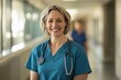© robertuzhbt89 - A smiling female healthcare professional wearing scrubs and a stethoscope, standing in a hospital corridor. The background shows blurred figures of other medical staff.