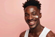 © SOLDATOOFF - An African American man applying cream on his face, smiling against a pink background