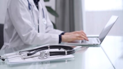 Tablet computer and black stethoscope are on the glass table besides a doctor woman using laptop computer on the glass table. Medicine concept