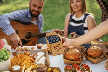 Guy is with acoustic guitar, having fun. Group of friends are having picnic on the field with food in eco boxes